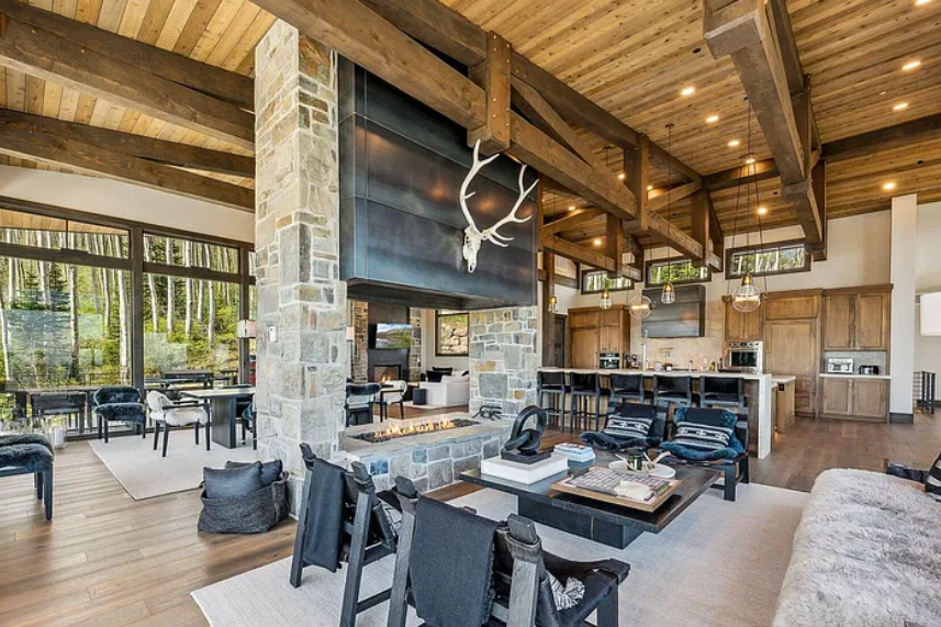 Spacious living room with stone fireplace, wood ceiling beams, large windows showing a forest view, and modern furniture.