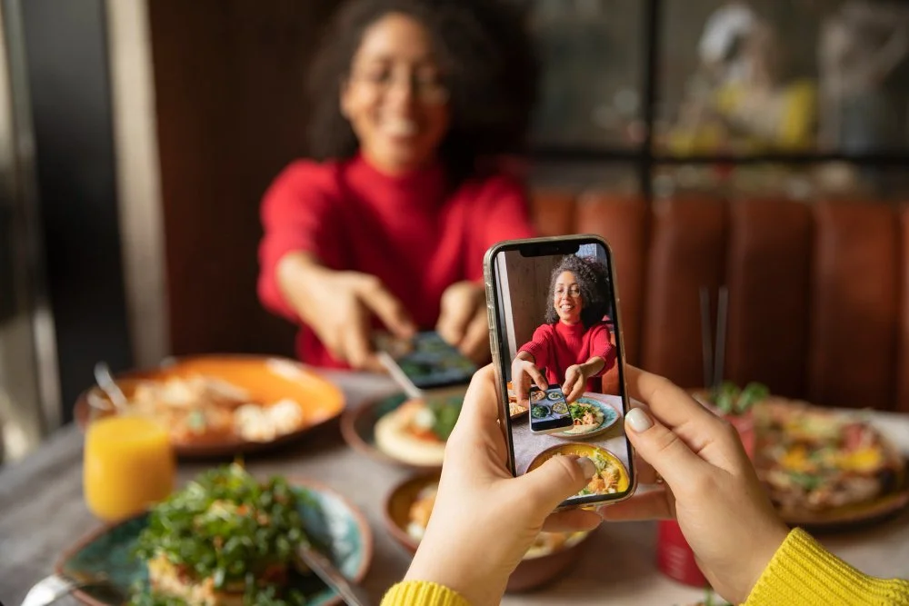 Person taking a photo of a woman with curly hair wearing a red shirt at a table with food, using a smartphone.
