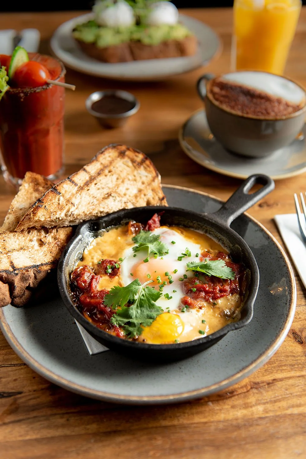 Fried egg in a small cast iron skillet with tomato sauce, garnished with cilantro and chives, served with toasted bread slices on a gray plate, with a cup of cappuccino and a glass of orange juice in the background.