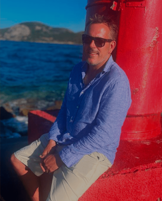 A man sitting outdoors near water with mountains in the background, wearing sunglasses, a blue button-up shirt, and light-colored shorts, leaning against a large red structure.