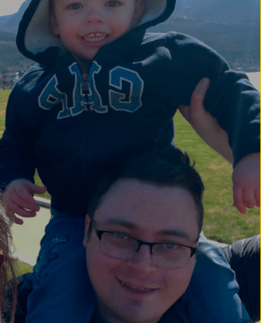 A man with glasses and short dark hair smiling, carrying a young boy on his shoulders outdoors on a sunny day, with green grass and trees in the background.