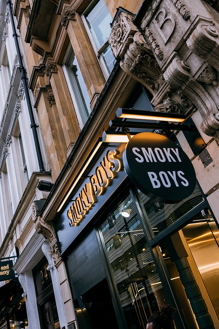 Street view showing a storefront with a sign reading 'Smoky Boys' and another sign with the text 'SERIOUS'. The building has ornate architectural details such as carved stone decorations around the windows and columns, with modern signage and lighting.