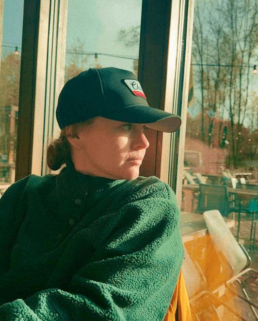 A woman sitting by a large window, wearing a black baseball cap and a green fleece jacket, looking outside.