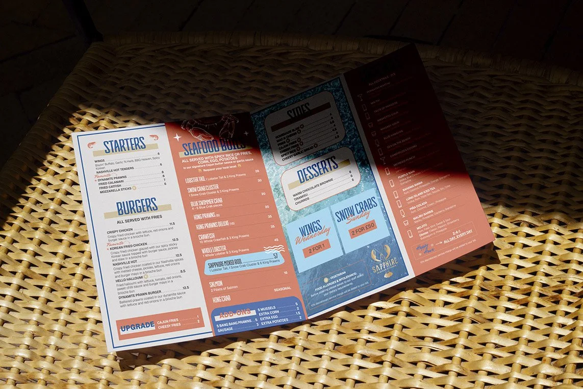 Restaurant menu placed on a wicker surface, featuring sections for starters, burgers, seafood boils, desserts, wings, snow crabs, and add-ons, with some items highlighted in red and blue.