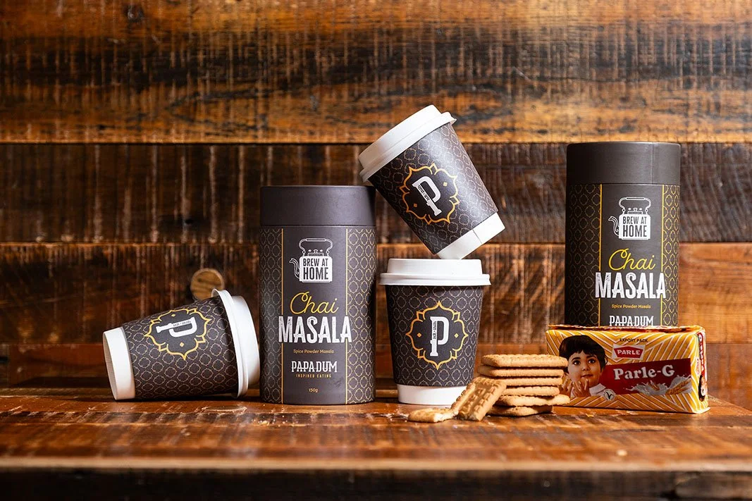 Various containers of Brew at Home Chai Masala spice with biscuits and a packet of Parle-G biscuits on a wooden surface against a wooden background.
