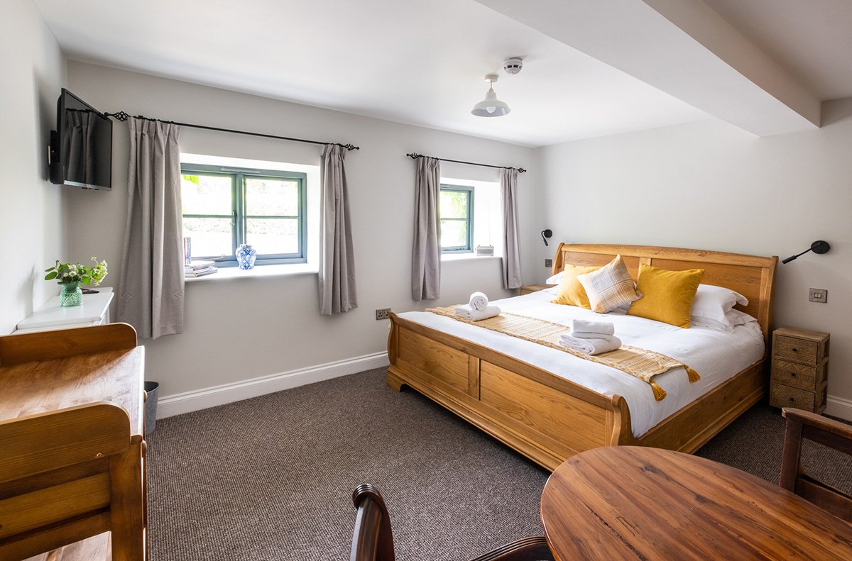 A bright bedroom with a large bed, two small windows with curtains, a wall-mounted TV, small side table, a desk with a plant, and chairs, decorated in neutral and wooden tones.
