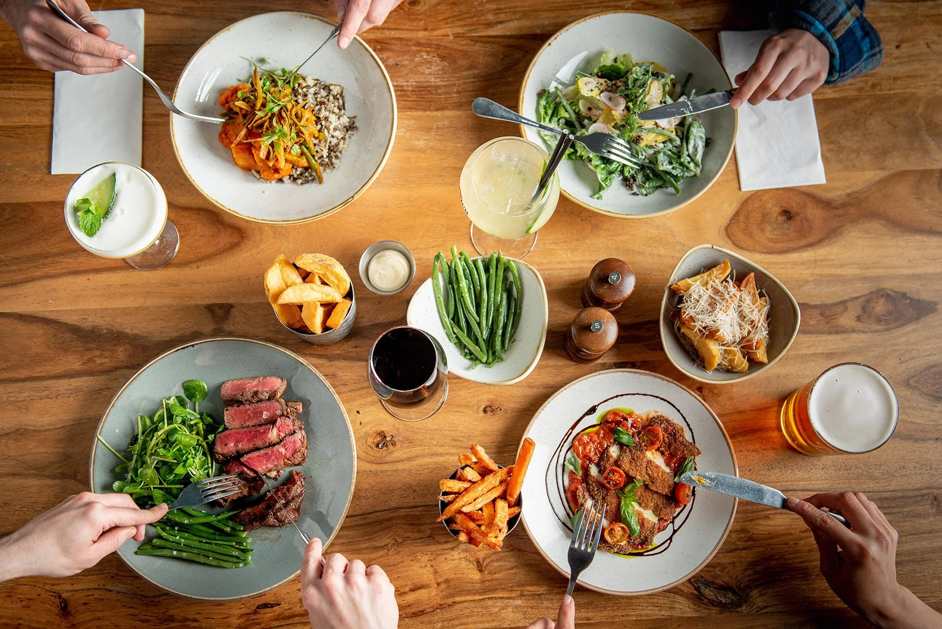 Overhead view of a wooden table with various plates of food, including steak, green beans, salad, pasta, and fried chips, with multiple hands using forks and knives.