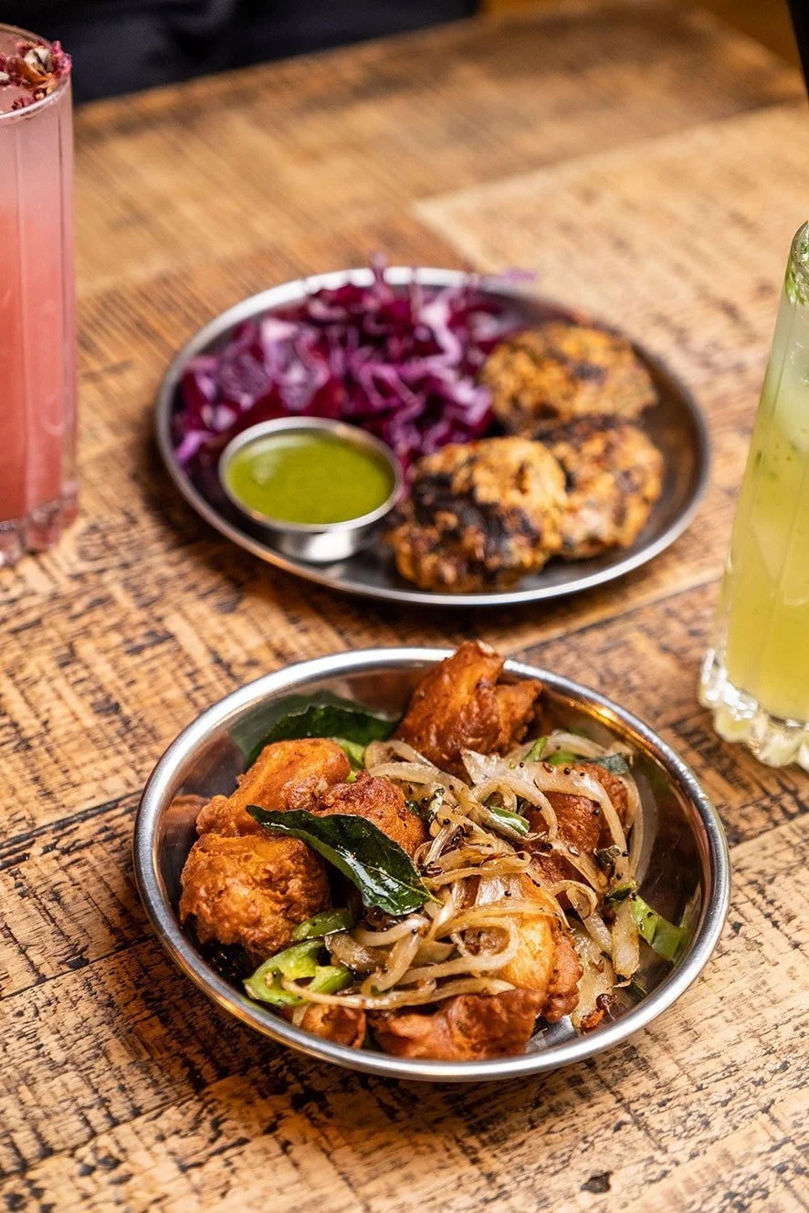 A wooden table with two bowls of Indian food; one containing fried chicken with sautéed onions and green chili, and the other with grilled meats, shredded purple cabbage, and green dipping sauce; two drinks are partially visible on the sides.