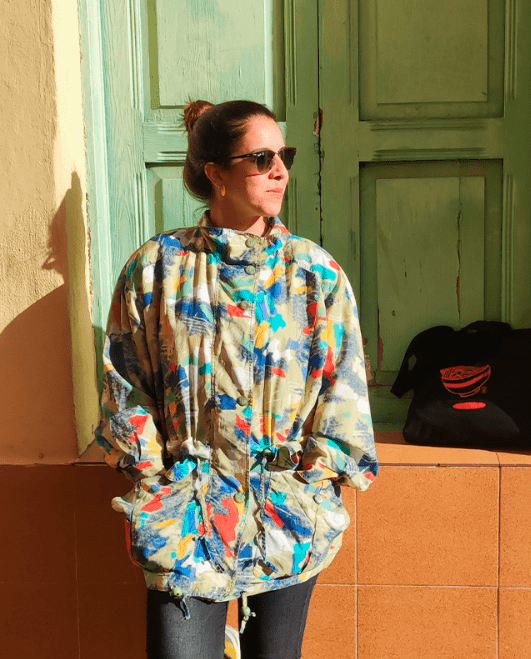 Woman wearing sunglasses and a colorful patterned jacket, standing against a yellow wall with green double doors, with a black bag on a ledge.