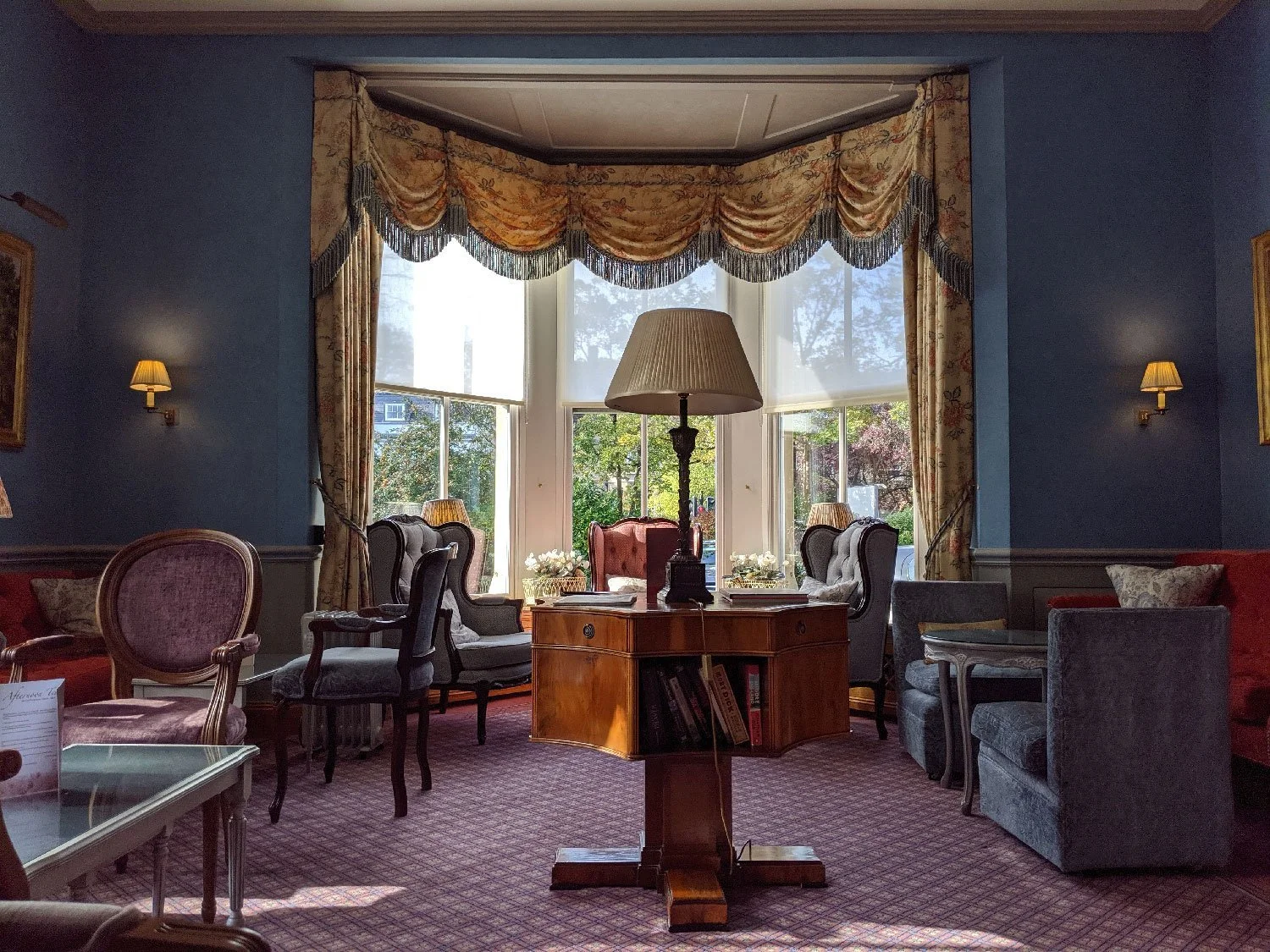 A cozy, elegant living room with a large bay window with floral and gold drapes. Inside, there is a central wooden side table with a large lamp, surrounded by vintage armchairs, sofas, and side tables. Outside the window, trees and houses are visible.