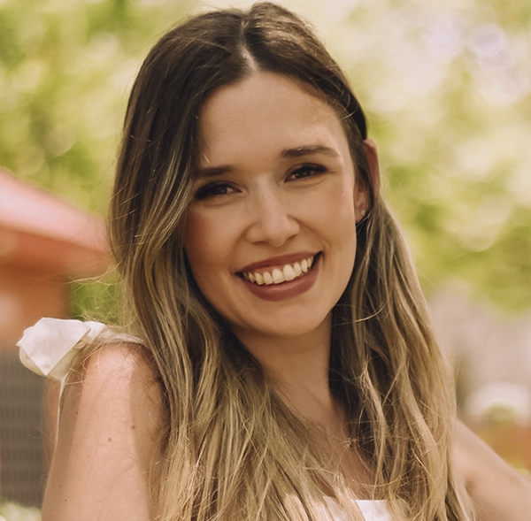 Young woman with long wavy hair smiling outdoors in sunny weather with green foliage background.