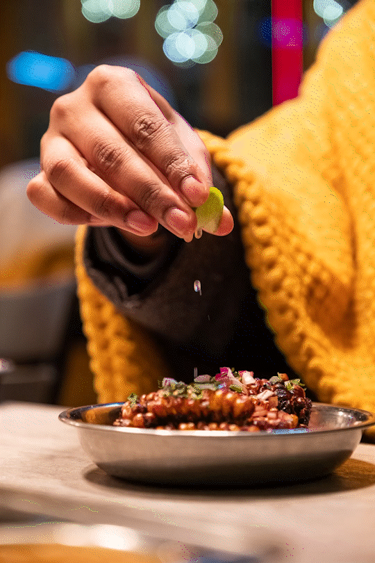 Restaurant marketing image of a person sprinkling lime juice onto a bowl of food, dressed in a yellow sweater, with colorful blurred lights in the background.