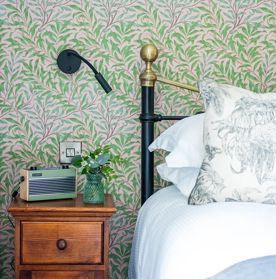 A bedroom with a wooden nightstand, a vintage radio, a green vase with plants, and a brass and black metal headboard. The wall behind features a pink and green leafy wallpaper, and a reading lamp is attached to the wall.