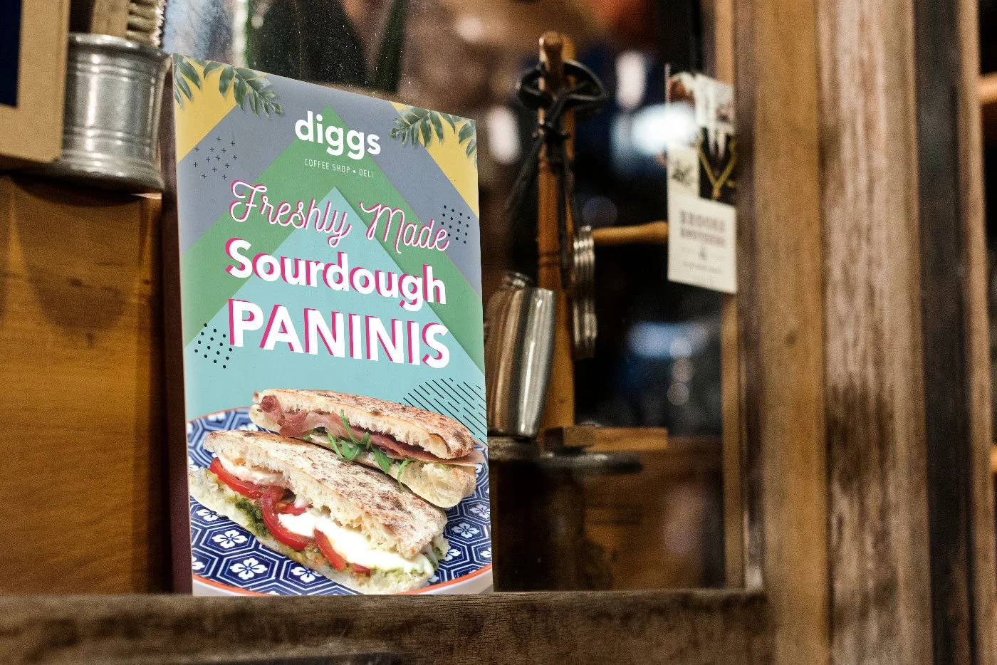 A colorful poster advertising freshly made sourdough paninis, with a picture of a panini sandwich filled with vegetables and cheese, on a plate with a blue pattern, inside a wooden cafe or shop.