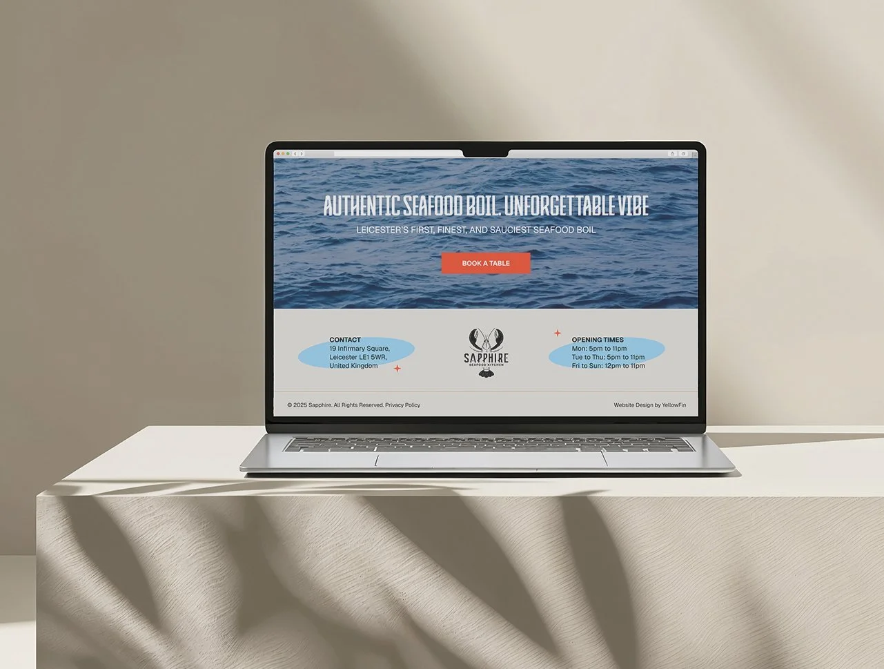Laptop displaying a seafood restaurant website with the headline 'Authentic Seafood Boil. Unforgettable Vibe' and a 'Book a Table' button, placed on a white table with shadows and a beige background.