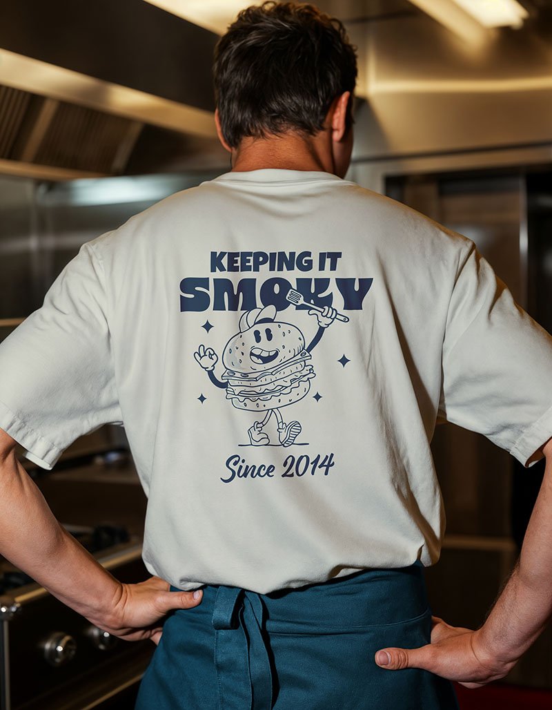 Restaurant Branding for Smoky Boys featuring the back of a person wearing a white T-shirt with a cartoon burger character, holding a fork, and the text 'Keeping It Smoky Since 2014' in a restaurant kitchen.
