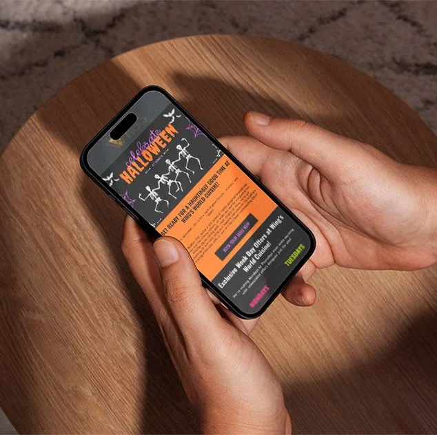 Person holding a smartphone displaying a Halloween event webpage, with a wooden table surface beneath.