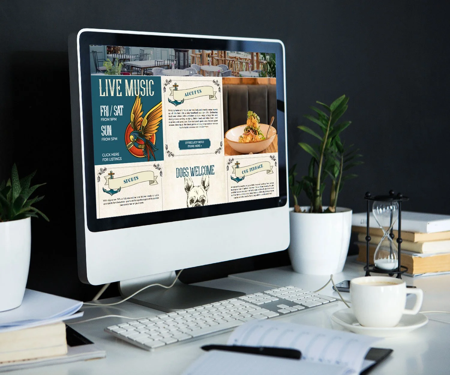 Computer monitor displaying a restaurant or cafe website with sections about live music, food, sports, and a dog welcome sign, surrounded by potted plants, a stack of books, a coffee cup, a small hourglass, and a keyboard on a white desk.