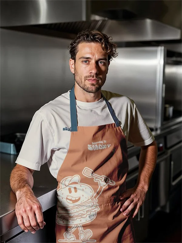 A man in a chef's kitchen wearing a beige apron with a cartoon burger character and the words 'Keeping it Sizzly' on it. He has short wavy hair and a beard, looking confidently at the camera.
