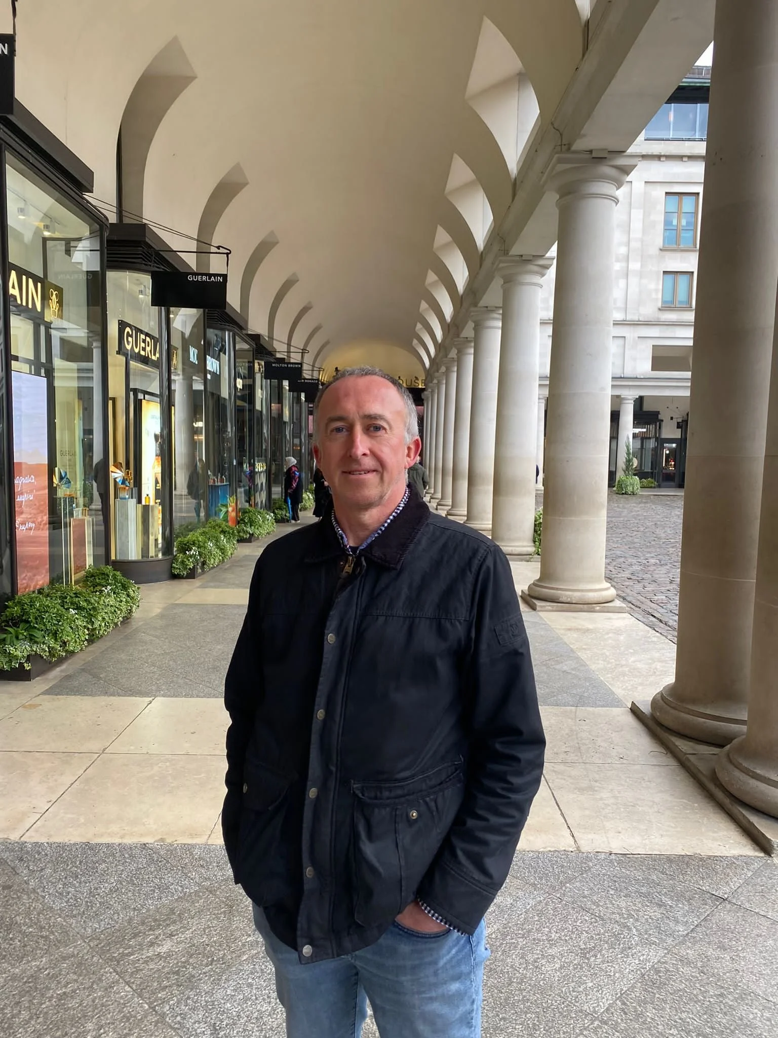 A man with short gray hair wearing a dark jacket and blue jeans standing under a covered walkway with white arches and columns, shopping windows with designer brand signs, and an urban building in the background.