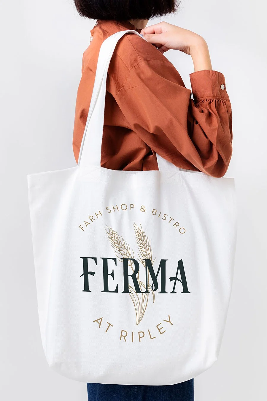 A woman holds a white tote bag with "FERMA" printed on it, along with "Farm Shop & Bistro at Tripley" and wheat illustrations.