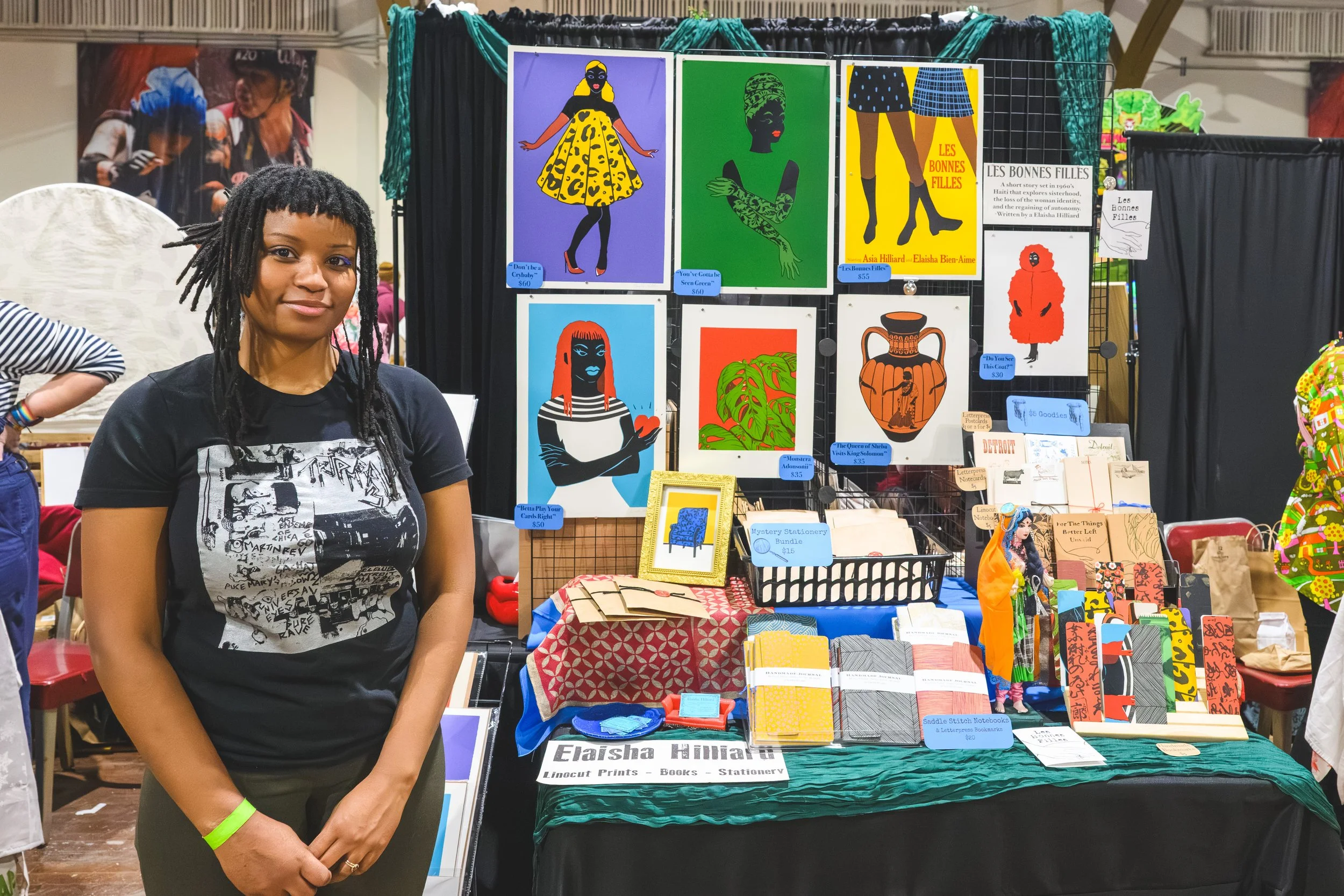 A artsy woman selling her beautiful artwork at the Detroit Urban Craft Fair. The booth displays colorful artwork, including illustrations of women, plants, and other designs, along with books and stationery items.