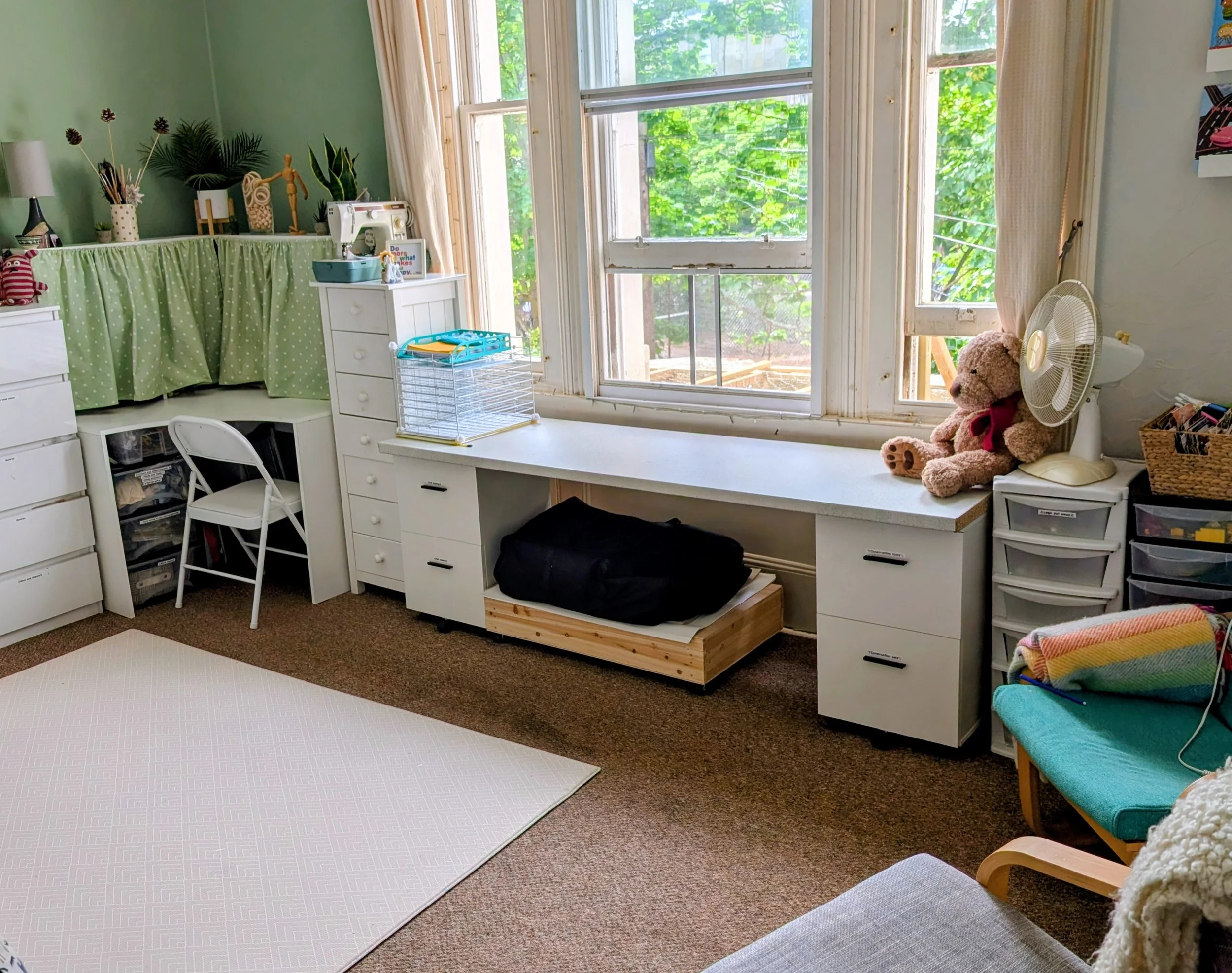 A room with large windows, white furniture, a teddy bear, a desk with a sewing machine, plants, and various decorations.