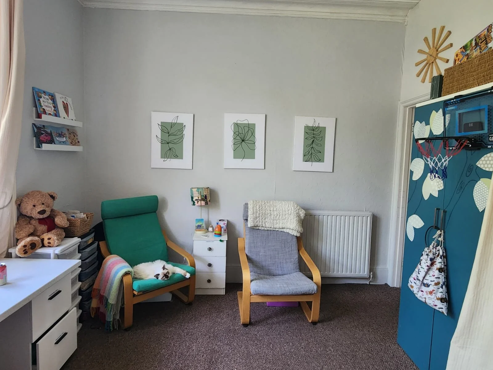 A cozy room with children's artwork on the wall, two chairs with blankets, a small white nightstand, toys, a teddy bear, a pile of books, a small white drawer unit, and a blue storage cabinet with a basketball hoop and toys hanging on it.