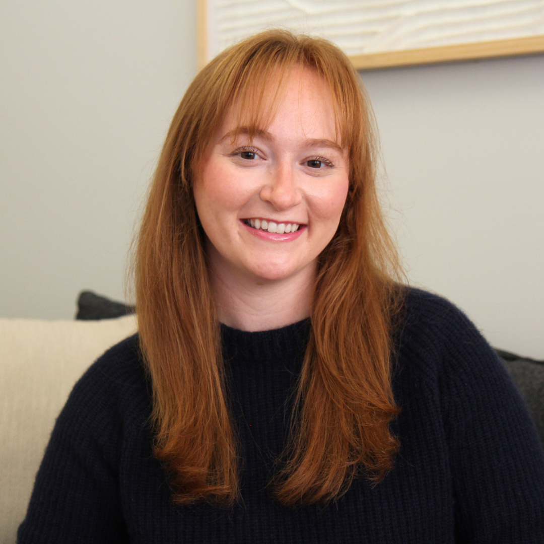 Smiling woman with long red hair wearing a black sweater, sitting on a cream-colored couch in a room with light-colored walls and a framed picture behind her.