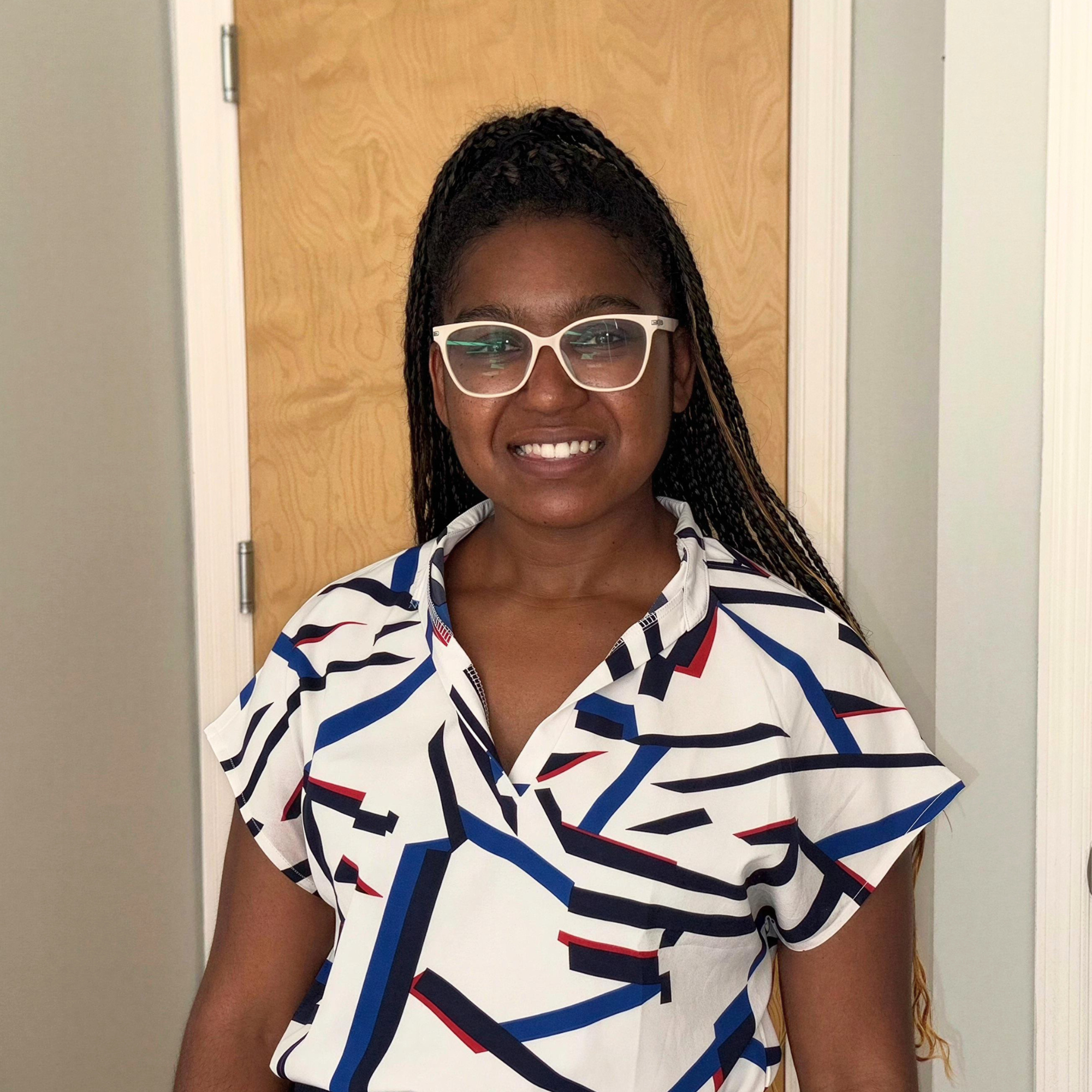 Smiling woman with glasses and braids, wearing a colorful geometric patterned blouse, standing in front of a wooden door.
