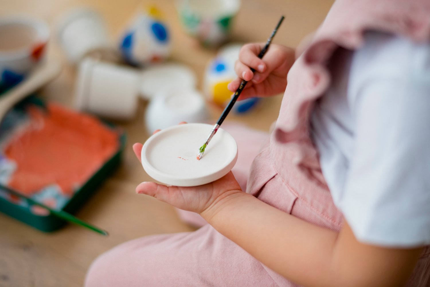 Pessoa pintando uma figura em uma tigela de cerâmica branca com um pincel fino. Ao fundo, há diversas tigelas de tinta colorida e uma bandeja com uma peça de artesanato em andamento.