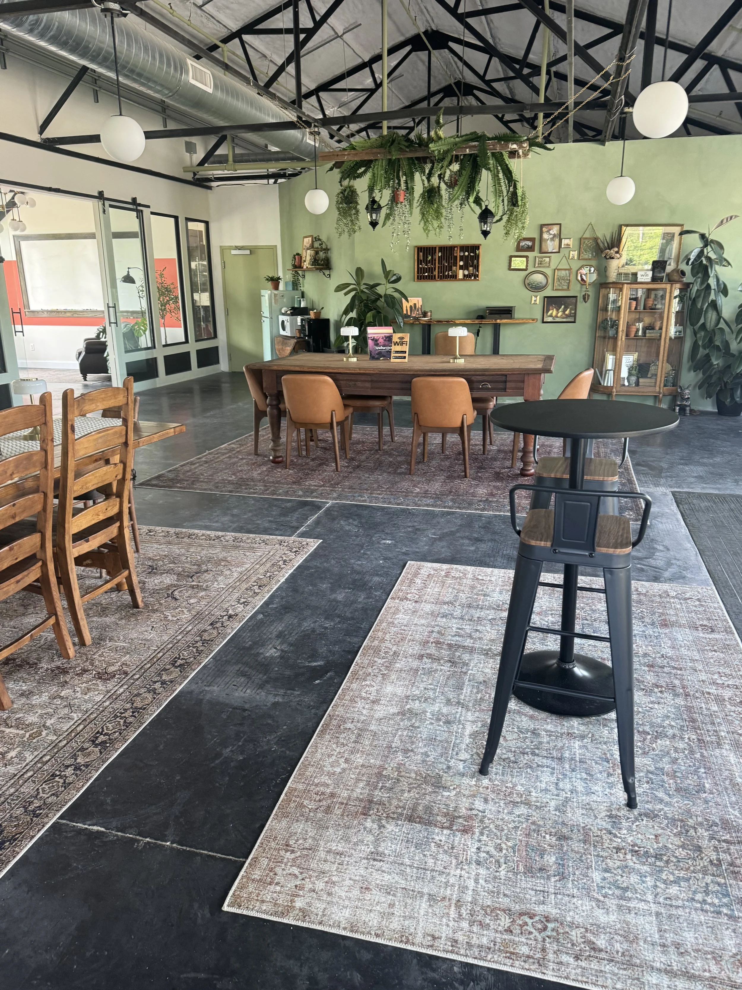 Interior of a cozy coffee shop with a mix of modern and rustic decor. The space features a long wooden table with tan chairs, a green accent wall decorated with framed pictures and plants, and hanging greenery from the ceiling. There are rugs on the dark floor, and various pieces of furniture, including a cabinet with glass doors and a counter with coffee equipment.