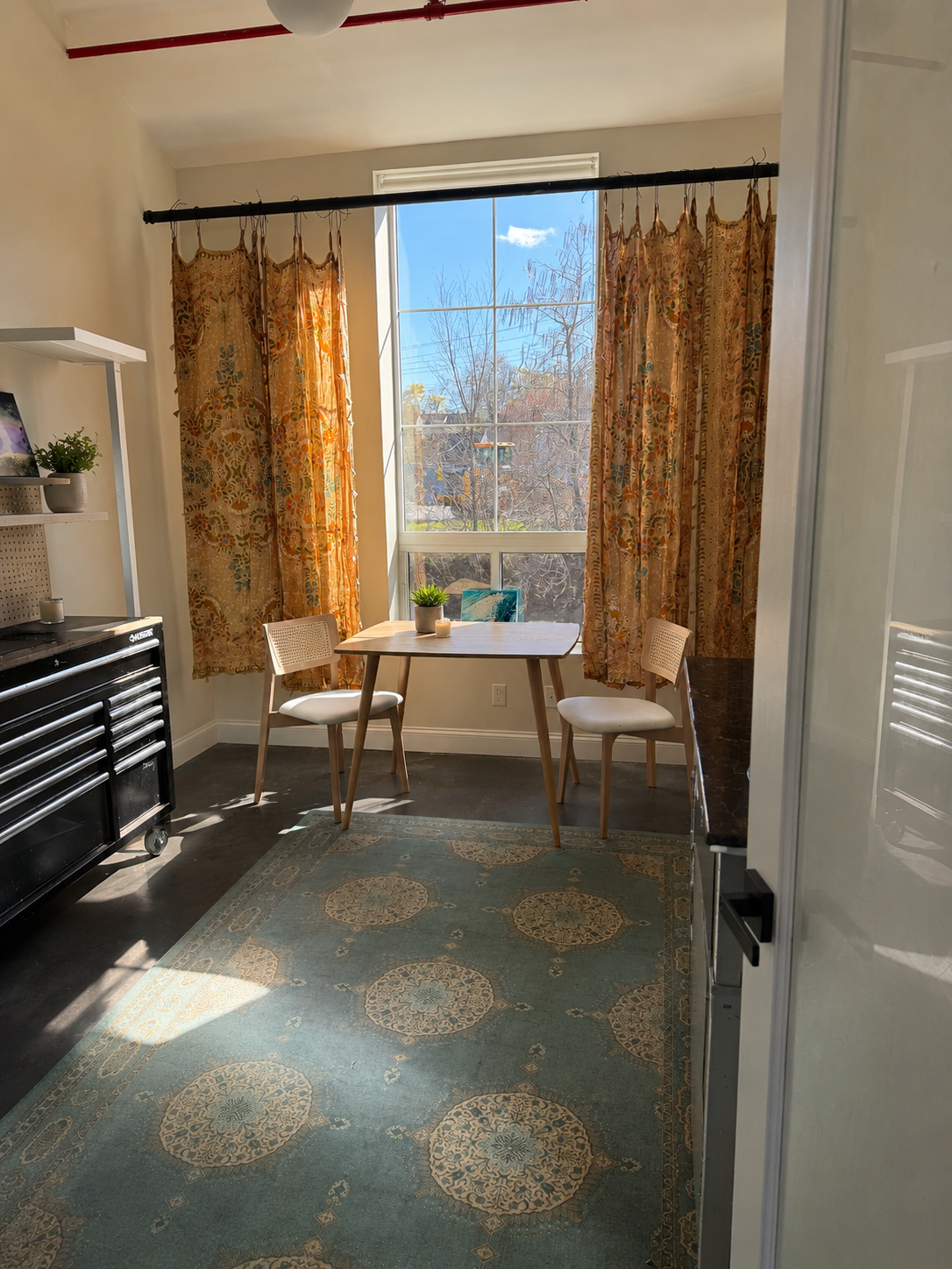 A cozy dining nook with a rectangular wooden table, two white chairs, and a large window with floral-patterned curtains, sunlight streaming in and casting shadows on the dark hardwood floor.