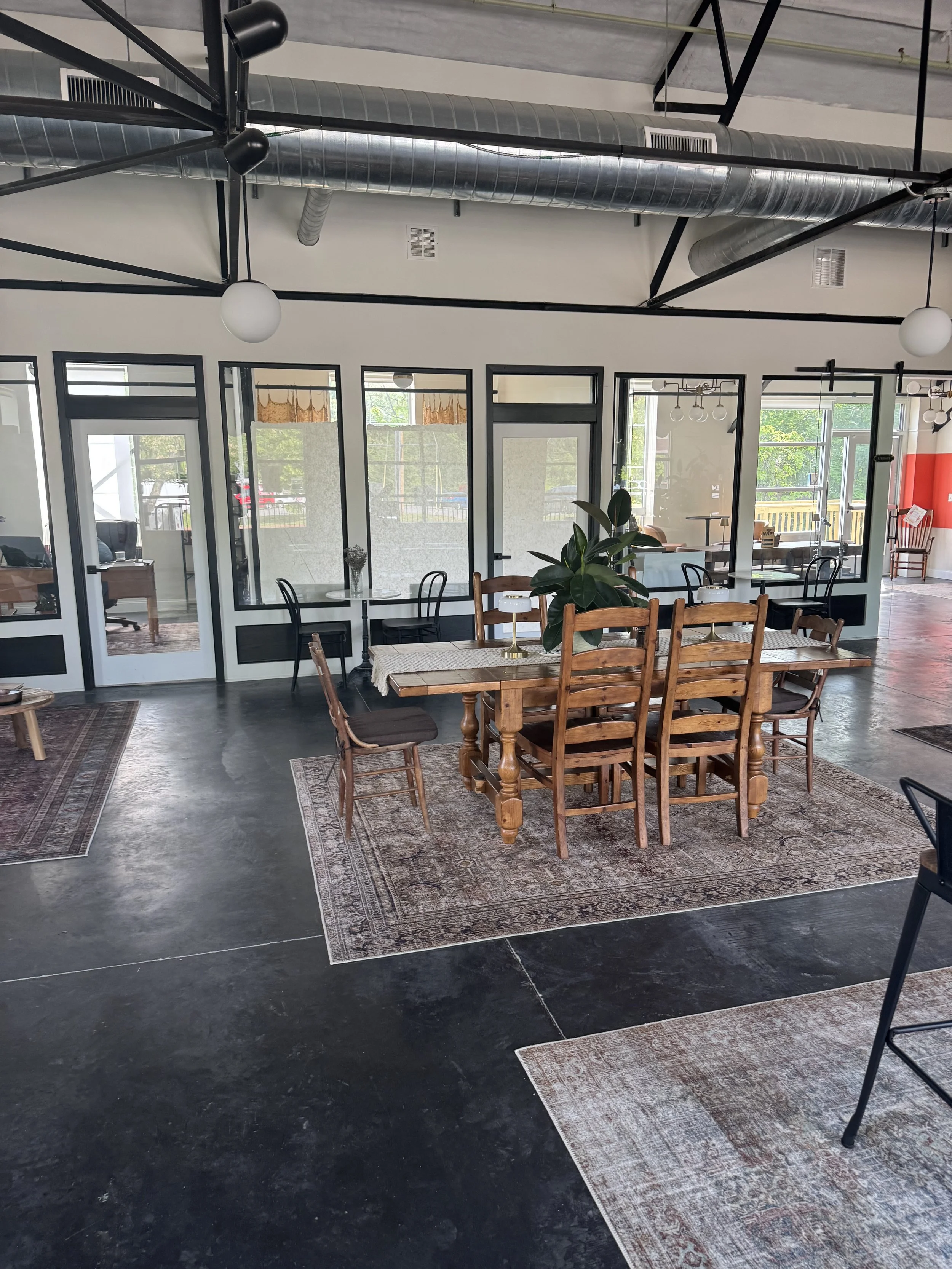 Interior of a modern cafe with a mix of wooden and metal furniture, large windows with black framing, a black polished concrete floor, exposed industrial ductwork on the ceiling, and decorative area rugs.