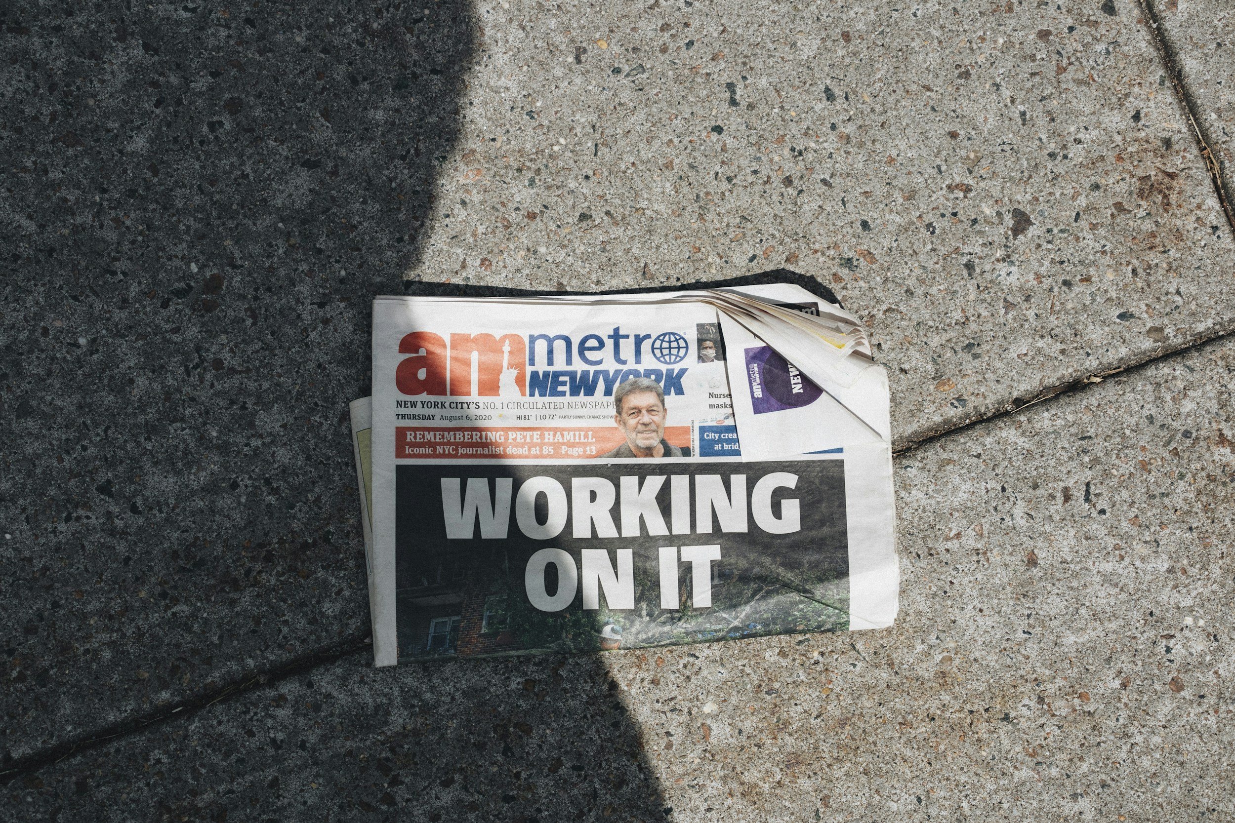 A crumpled newspaper lying on a concrete sidewalk with its headline partially visible, reading "WORKING ON IT."