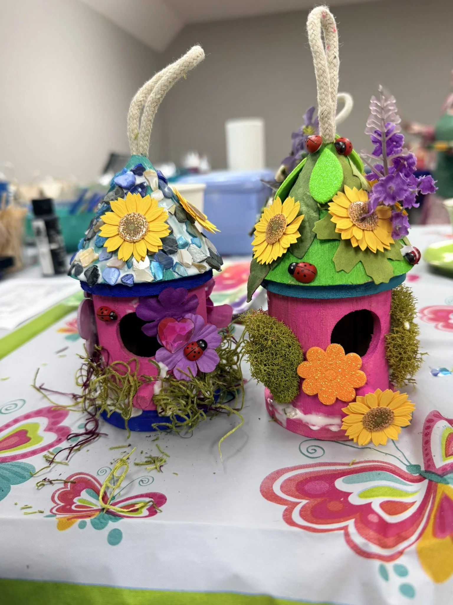 Colorful craft fairy houses decorated with flowers, ladybugs, and moss, placed on a table with a vibrant tablecloth featuring butterfly designs.