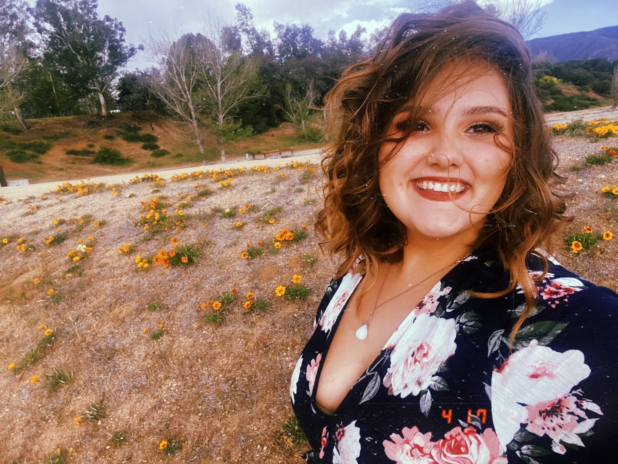A woman with curly brown hair smiling outdoors in a field of small yellow flowers, with trees and mountains in the background, wearing a floral patterned dress.