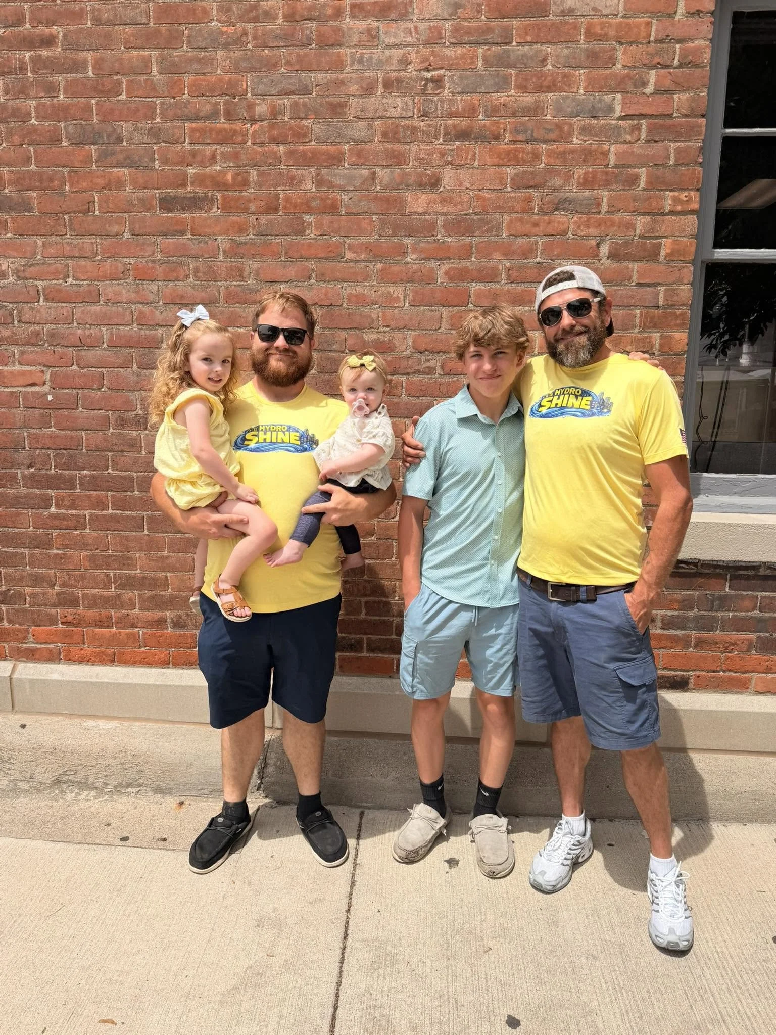 Five people, two adult men, one young man, and two young girls, standing in front of a brick wall. The men are wearing yellow t-shirts with a logo that says 'Hydro Shine,' and the young man is wearing a light green shirt. The girls have bows in their hair, and the group appears to be enjoying a sunny day outside.