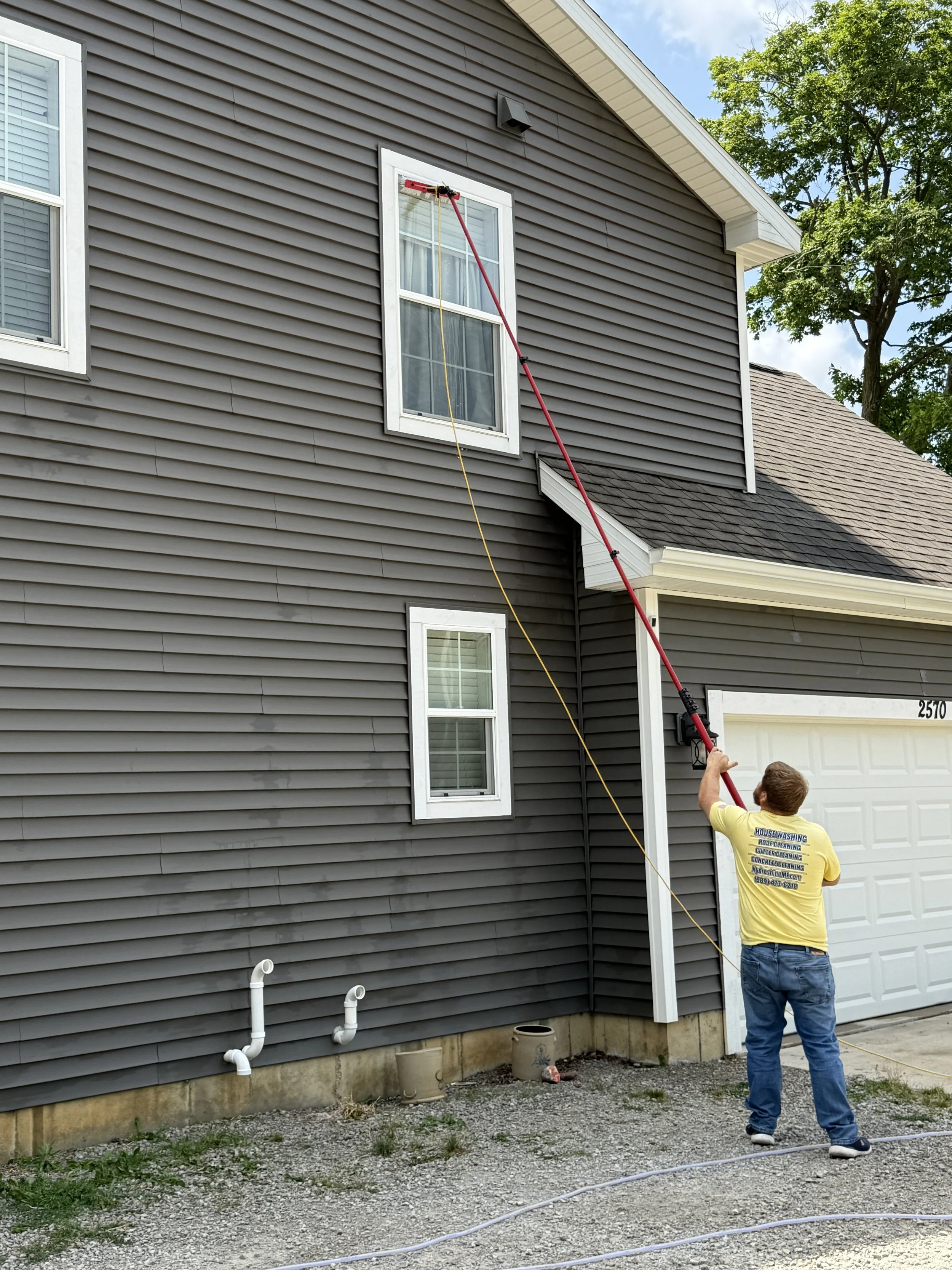 A man is pressure washing the side of a house with gray vinyl siding. The man is wearing a yellow shirt and jeans, holding a long red pressure washer extension arm. The house has white-framed windows, a garage door, and some white pipes on the ground nearby. Trees are visible in the background.