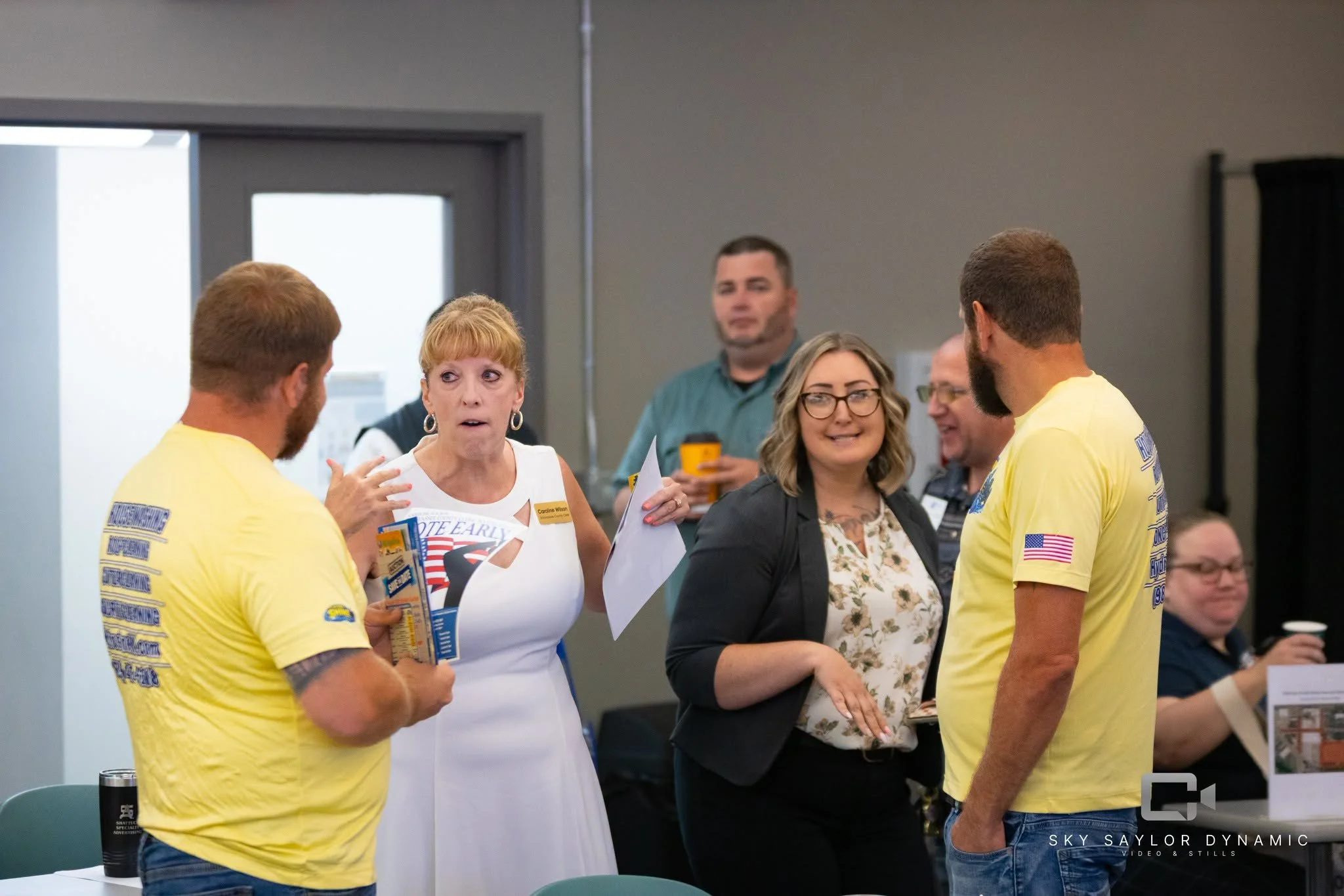 A group of people, including women and men, are engaged in conversation at an indoor event. Some individuals are wearing yellow T-shirts with text and American flag patches. One woman is holding papers, and other people are holding drinks. The settin