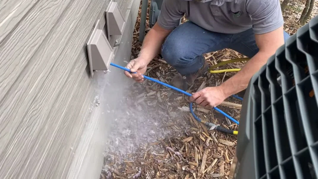 A person is pressure washing the exterior wall of a building near electrical outlets, with debris on the ground and outdoor equipment nearby.