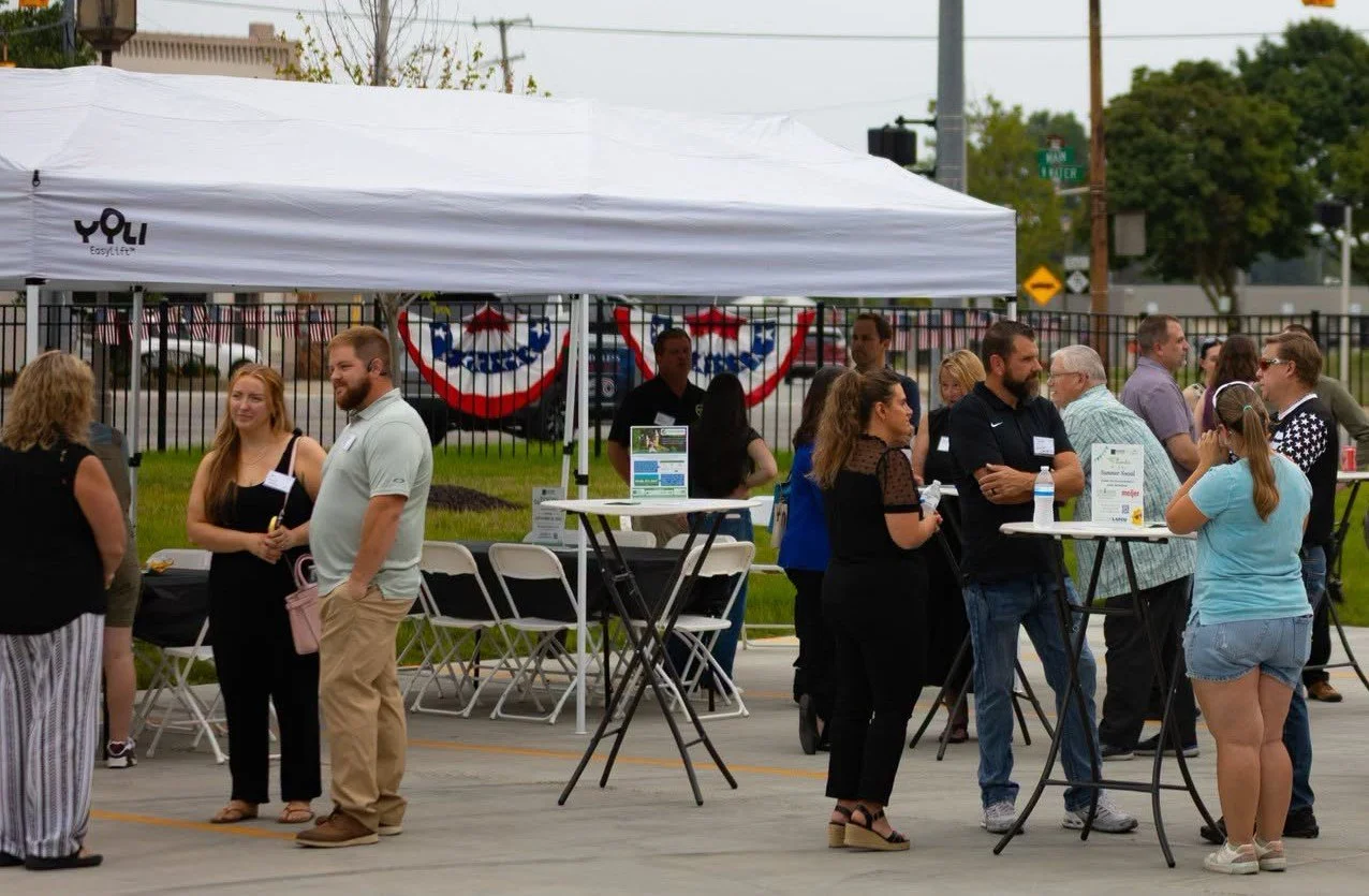 People socializing at an outdoor event with a white tent, tables, and patriotic decorations along a fenced sidewalk.
