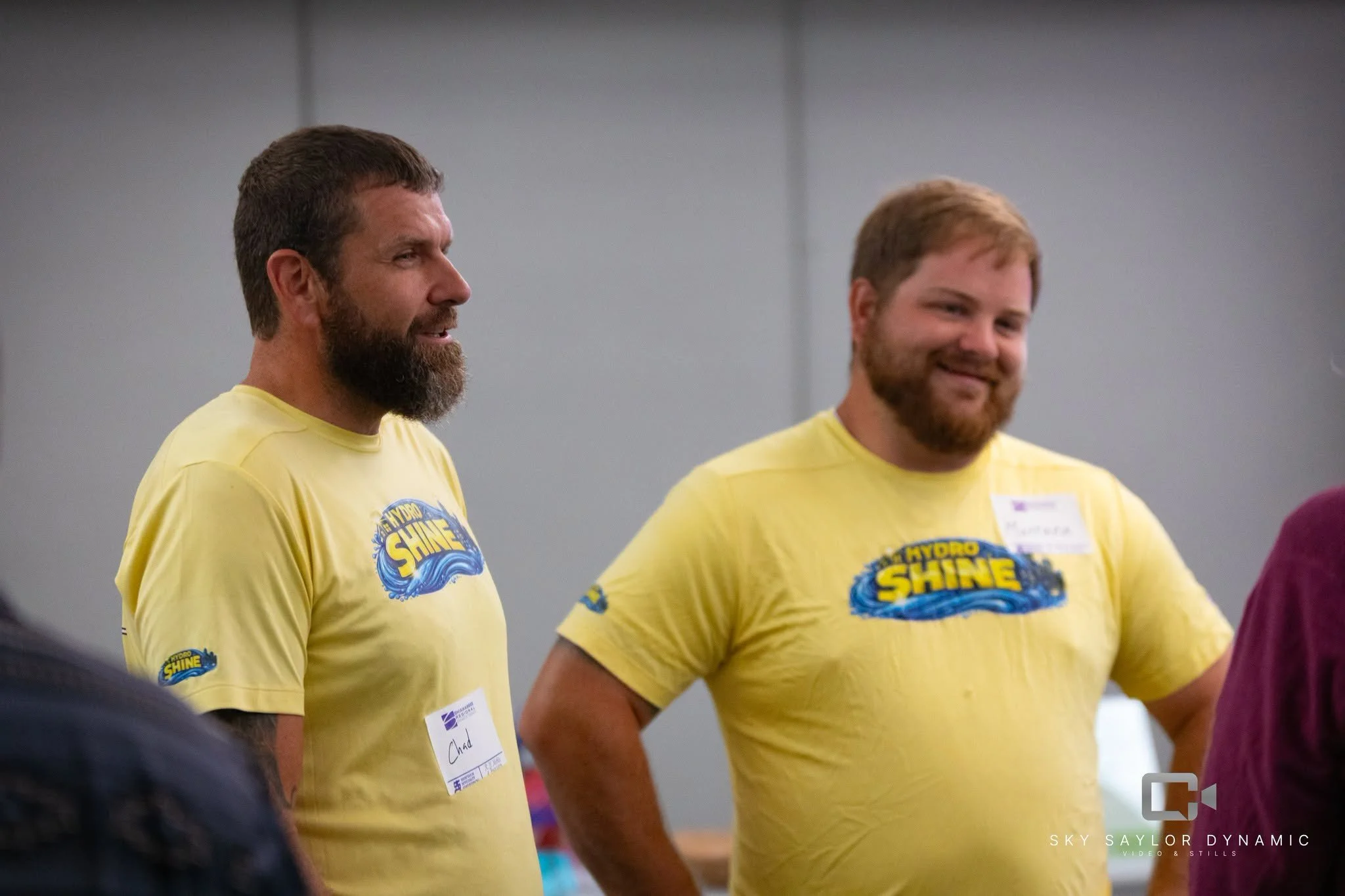 Two men wearing yellow T-shirts with a logo that says "Hydro Shine" standing indoors, smiling and talking to people.