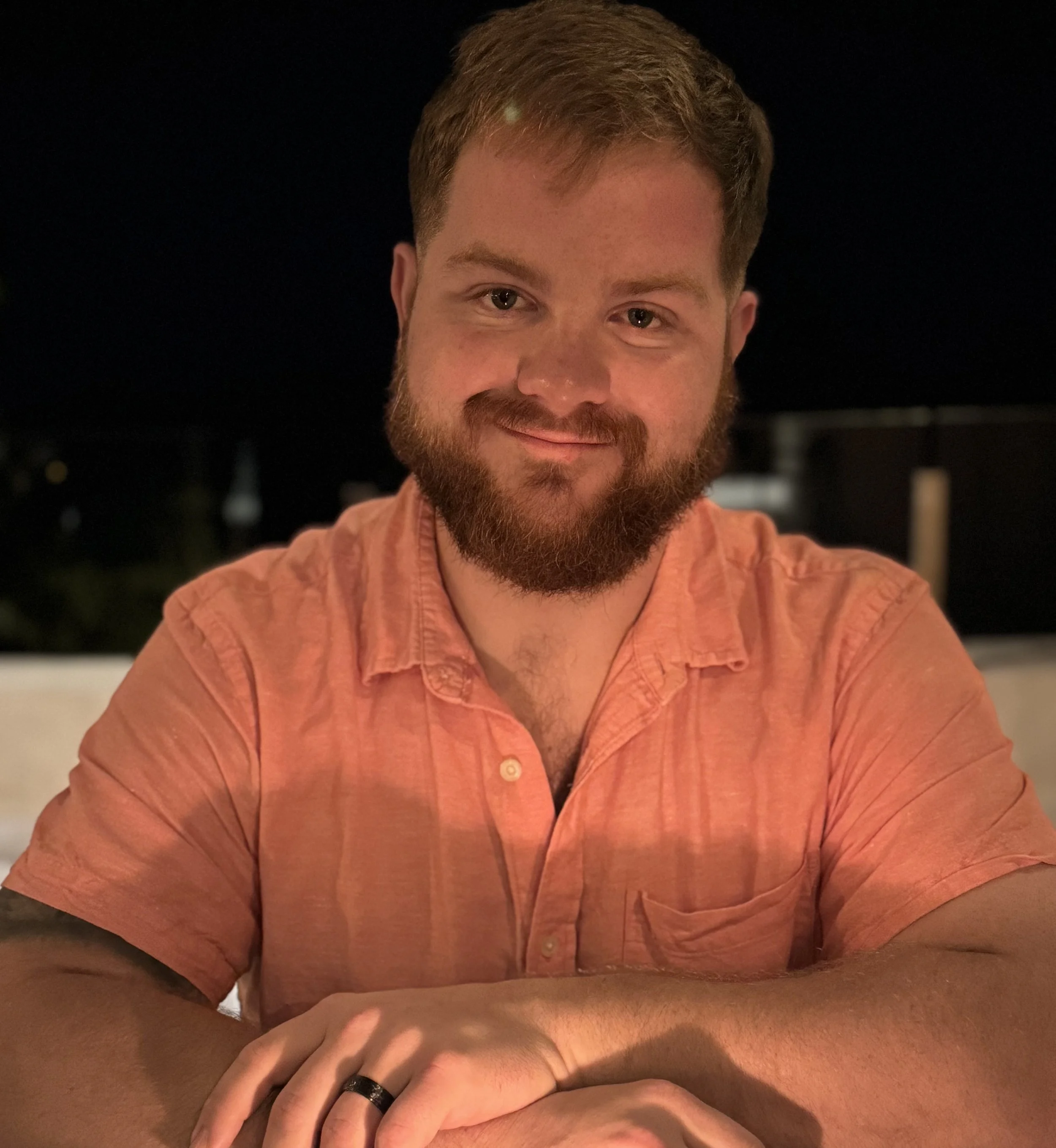Close-up of a smiling man with a beard and mustache, wearing an orange shirt, with his arms resting on a table, in a dimly lit environment.
