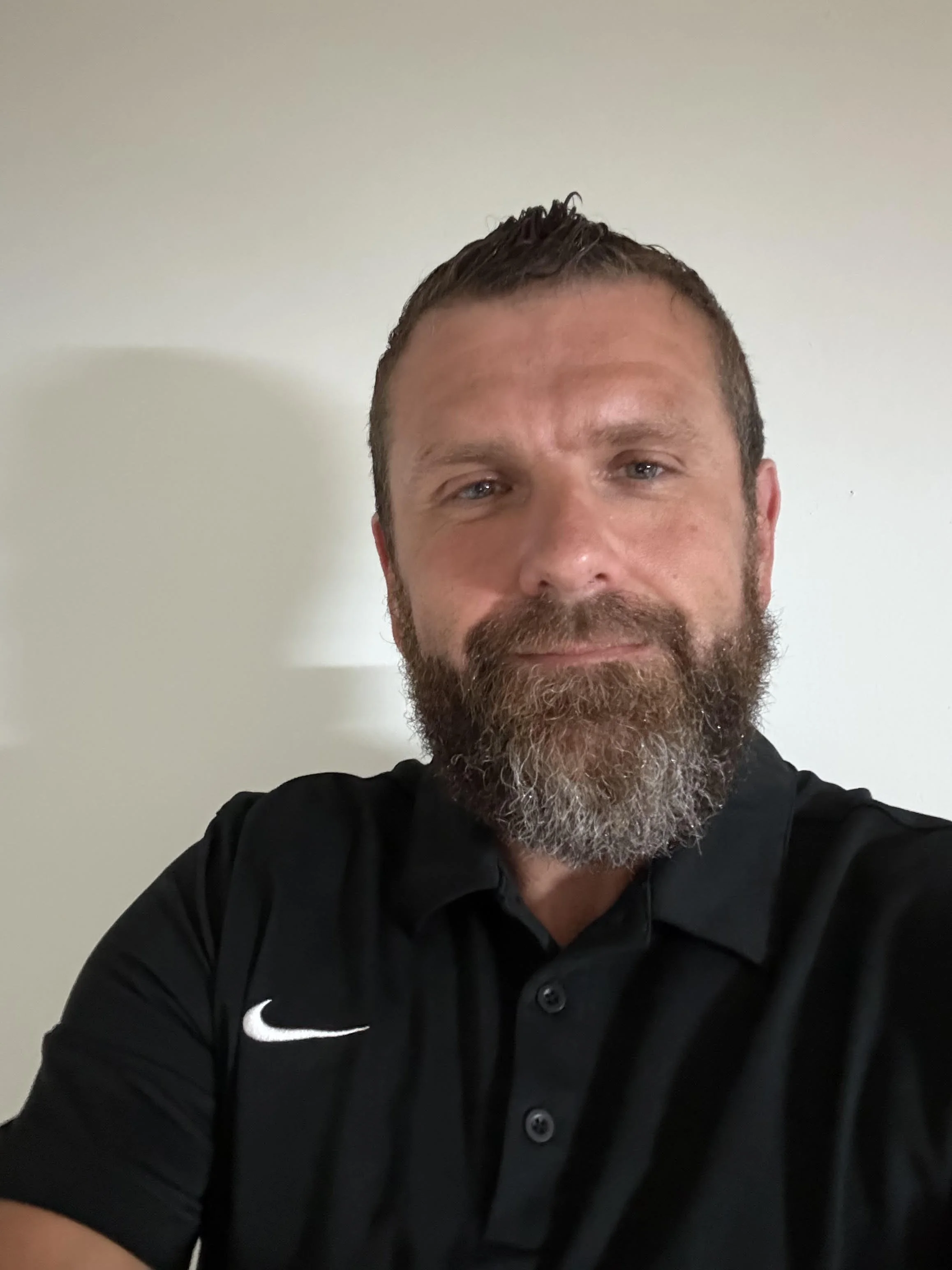 Close-up of a man with short dark hair, a beard, and blue eyes smiling, wearing a black Nike polo shirt, against a plain light-colored wall.