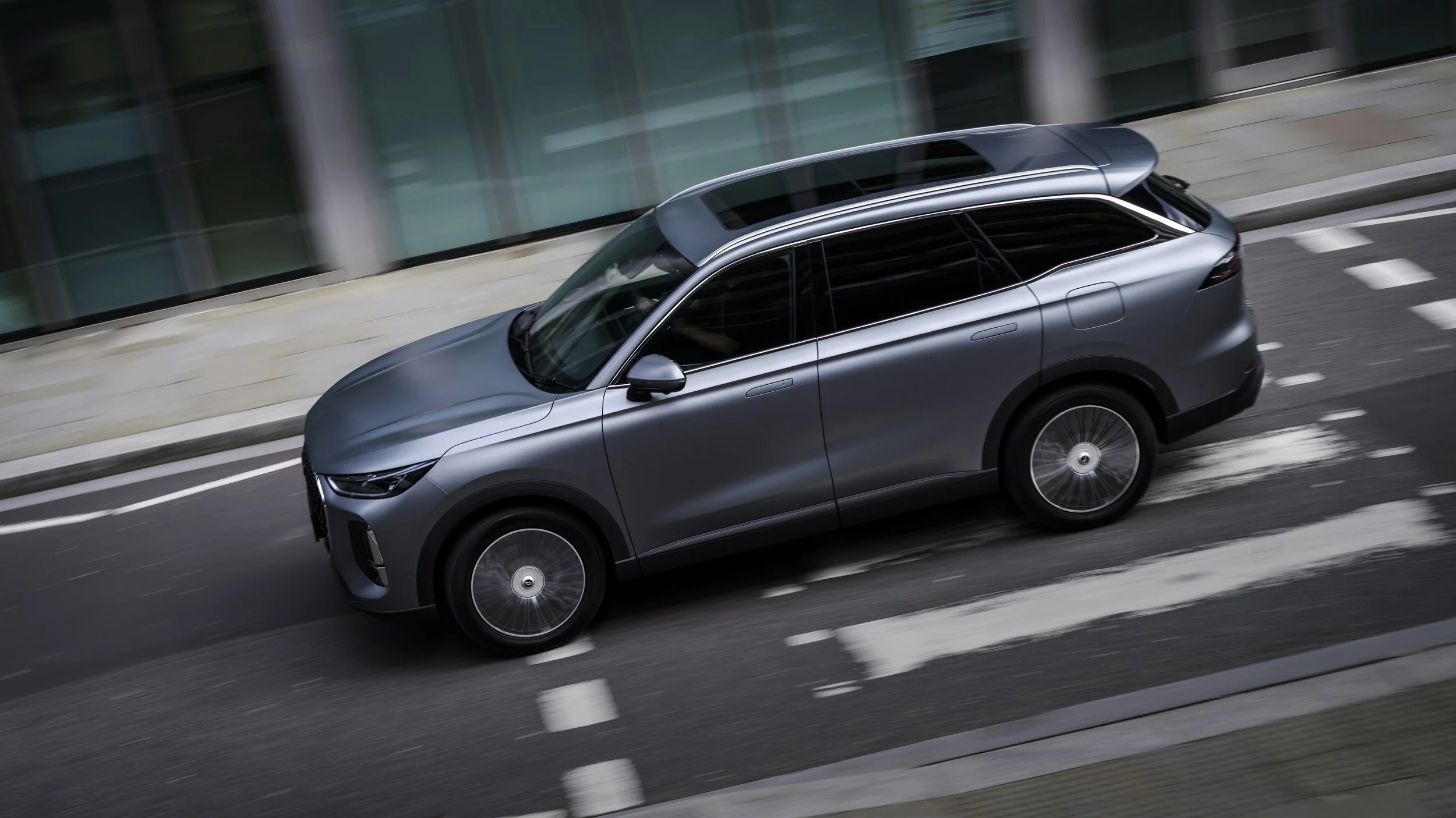 Silver-gray SUV driving on city street with glass building in background.