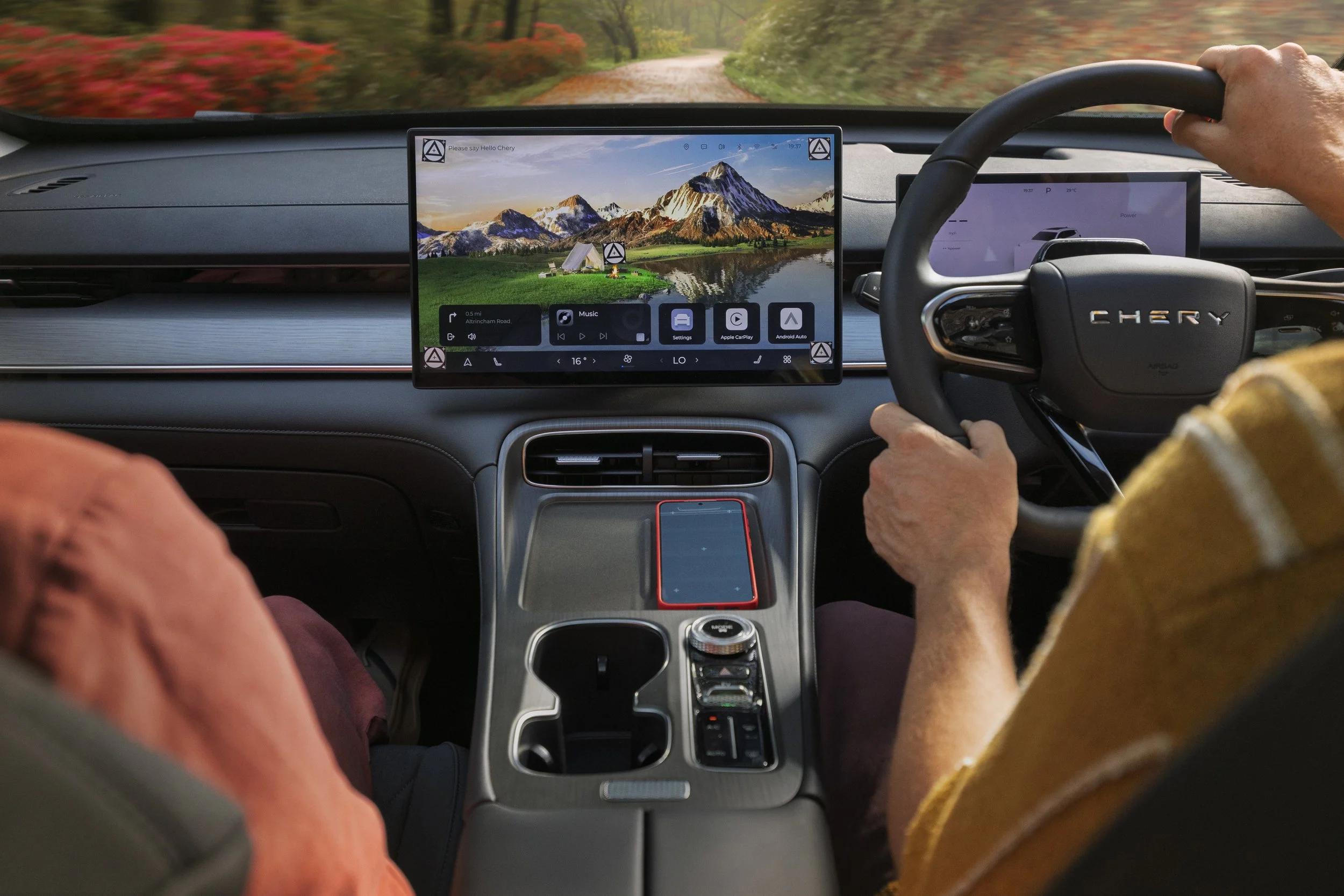 The interior of a car showing a dashboard with a large touchscreen, a steering wheel with 'CHERY' branding, and two passengers, one in a yellow striped shirt and the other in an orange shirt. Outside the windshield, a scenic mountain landscape with t