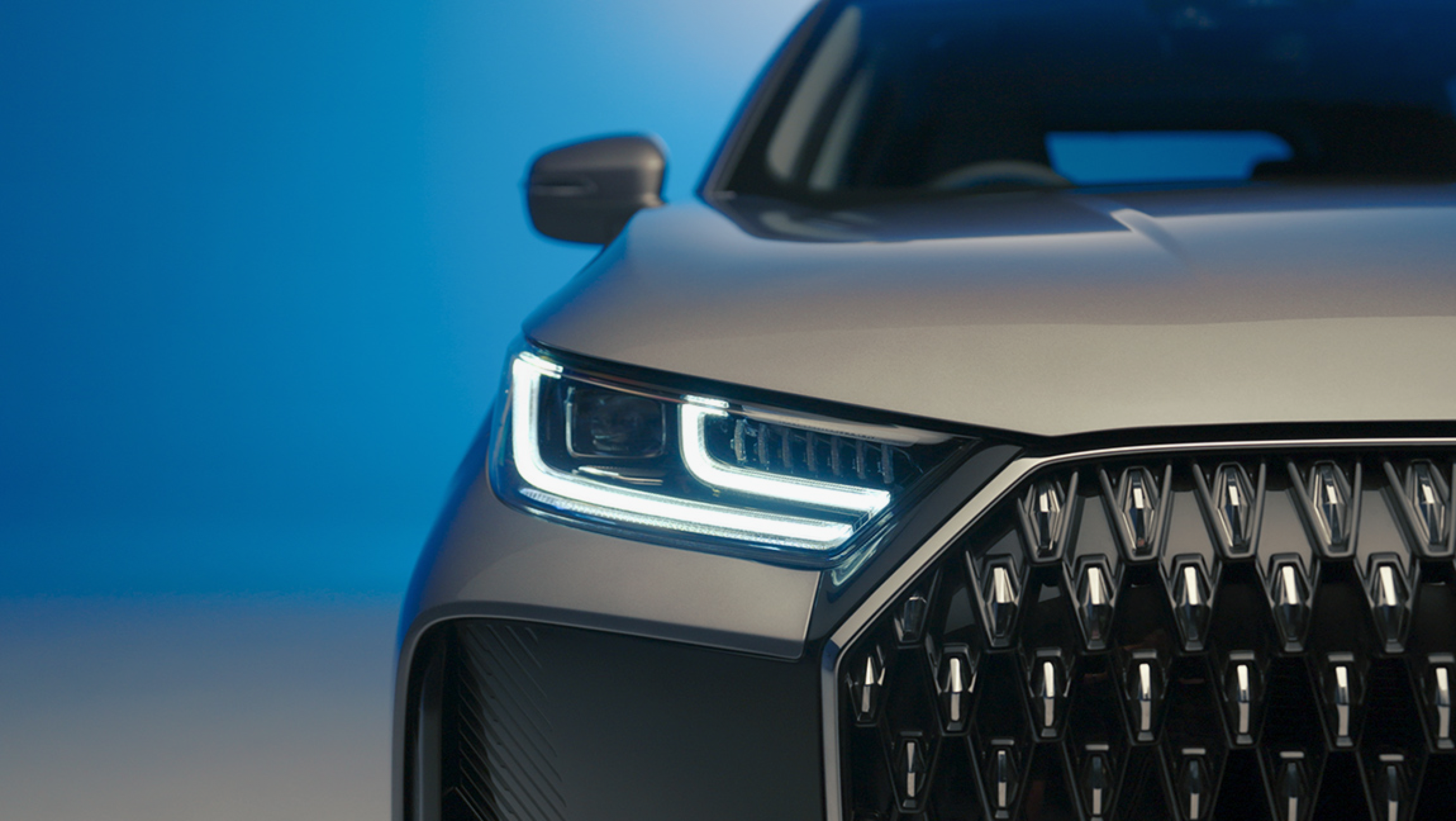 Close-up of the front left side of a silver modern SUV, showing the headlight, part of the grille, and side mirror against a blue background.
