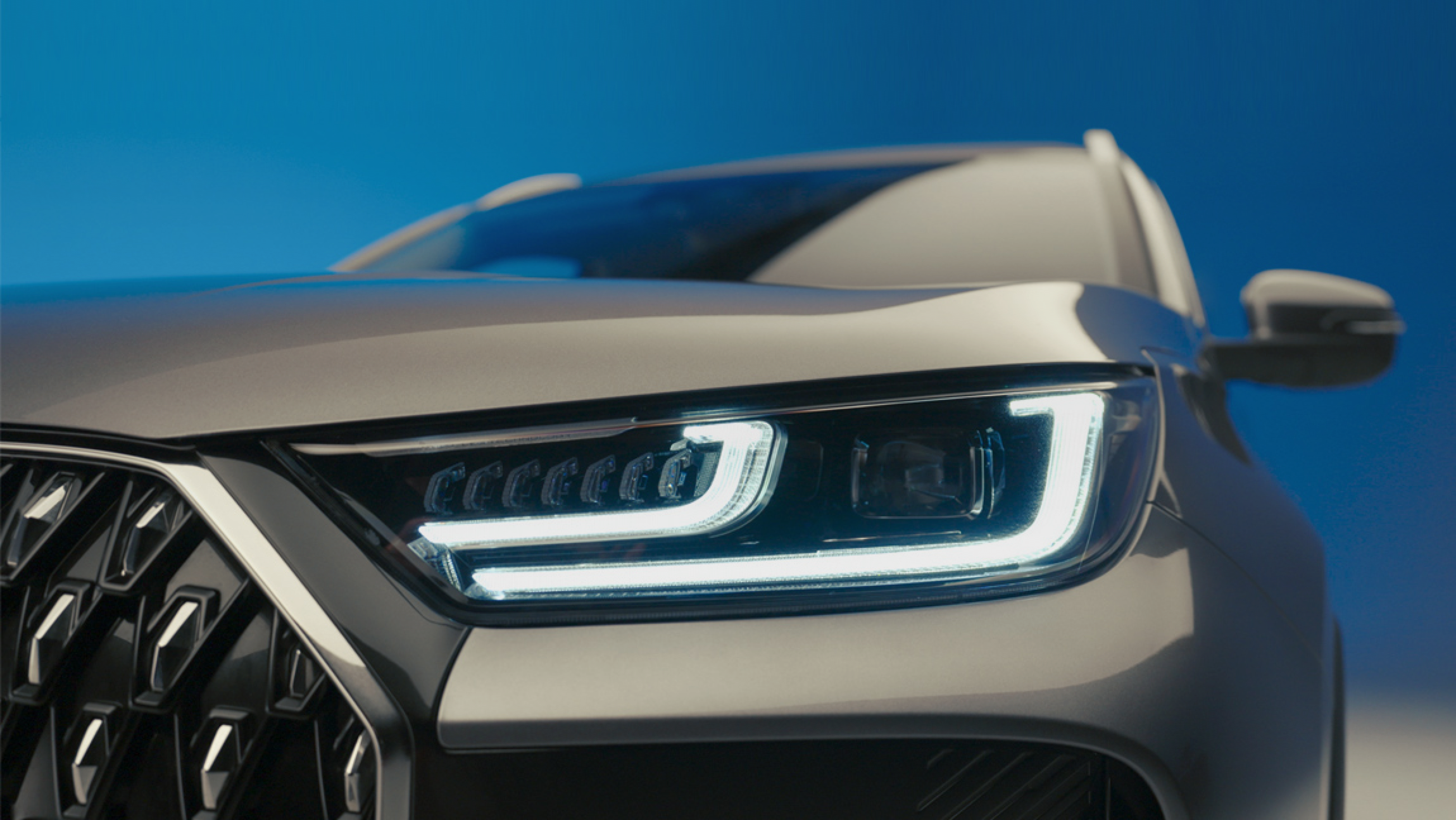 Close-up of a silver modern car's front with illuminated LED headlights against a blue sky background.