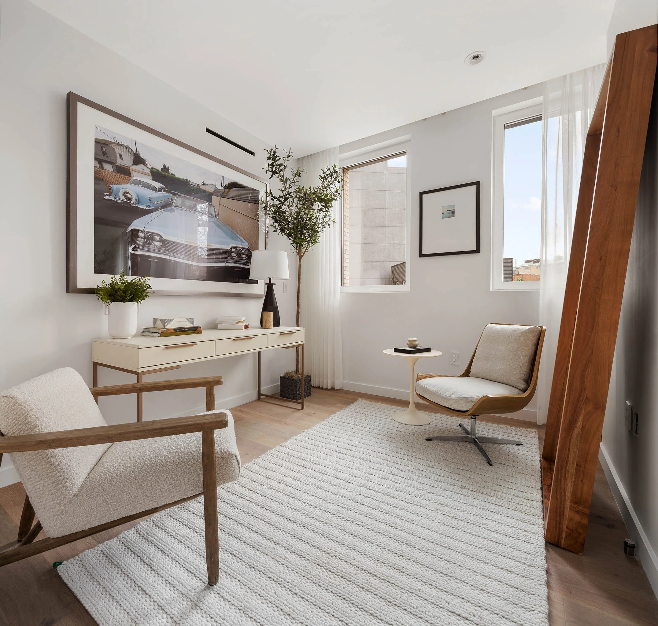 Bright, modern living room with white walls, wooden flooring, large windows with sheer curtains, contemporary furniture including a beige armchair, a white side table, and a white console table decorated with books, plants, and a black table lamp, wi